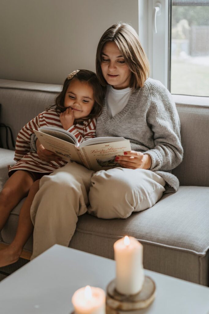 A cozy moment of a mother reading a book to her curious daughter on a couch.