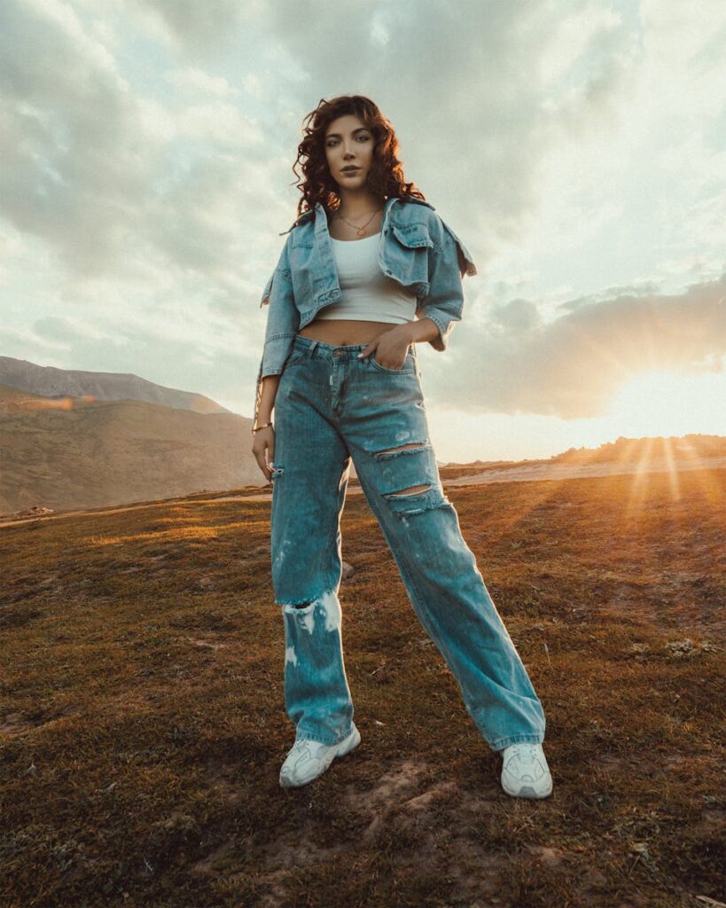 Full length of stylish female in denim outfit holding hand in pocket while standing on grassy field and looking at camera