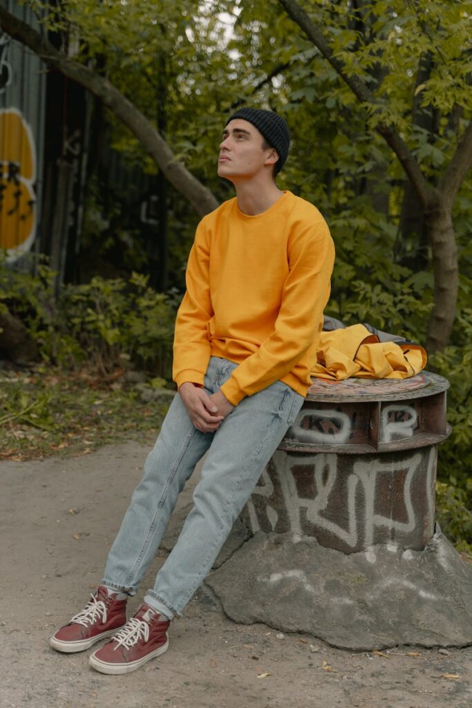 Young man in a yellow sweatshirt and beanie sitting outdoors, deep in thought.