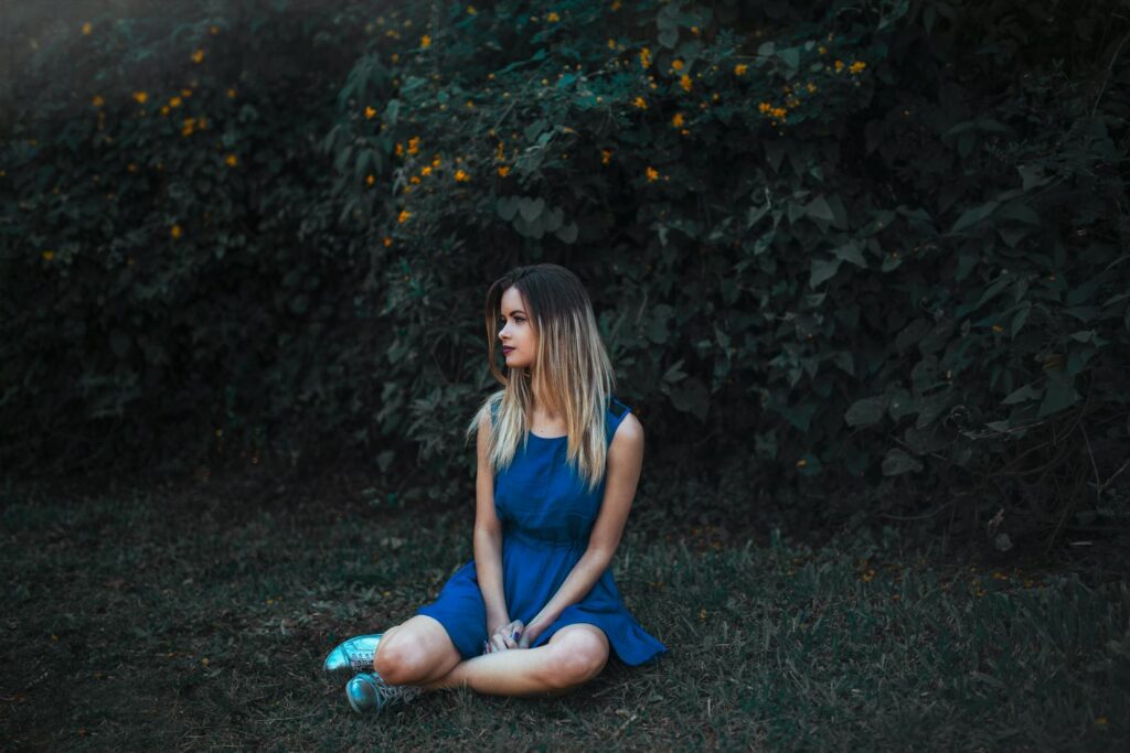 A young woman in a blue dress sits on the grass surrounded by lush foliage.