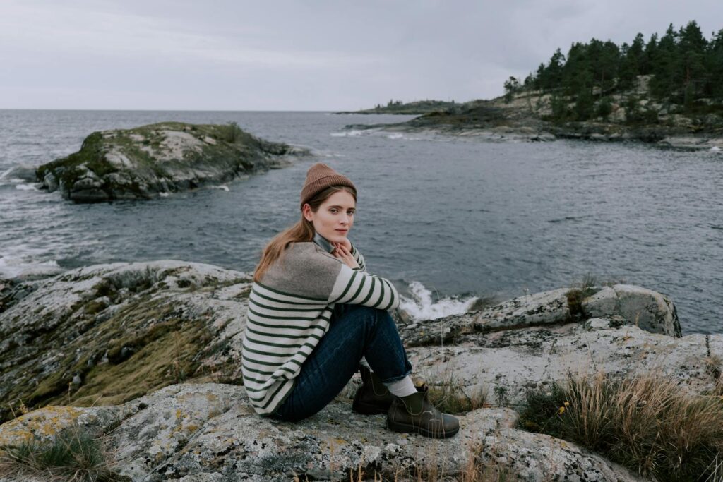 A woman in casual attire sits on a rocky shore by the sea, enjoying the serene nature.