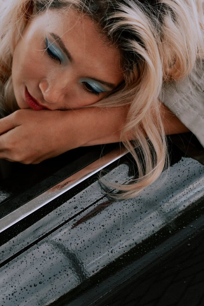 Close-up of a woman with blonde hair, blue eye makeup, and red lips resting on a reflective surface.