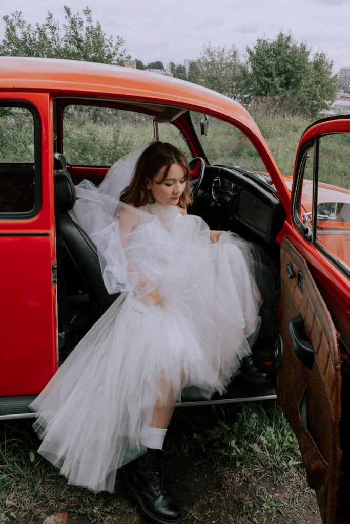 Young bride in a white wedding gown sitting in a vintage red car outdoors.