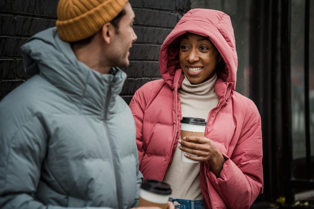Smiling couple in winter coats enjoying hot coffee outdoors on a chilly day.