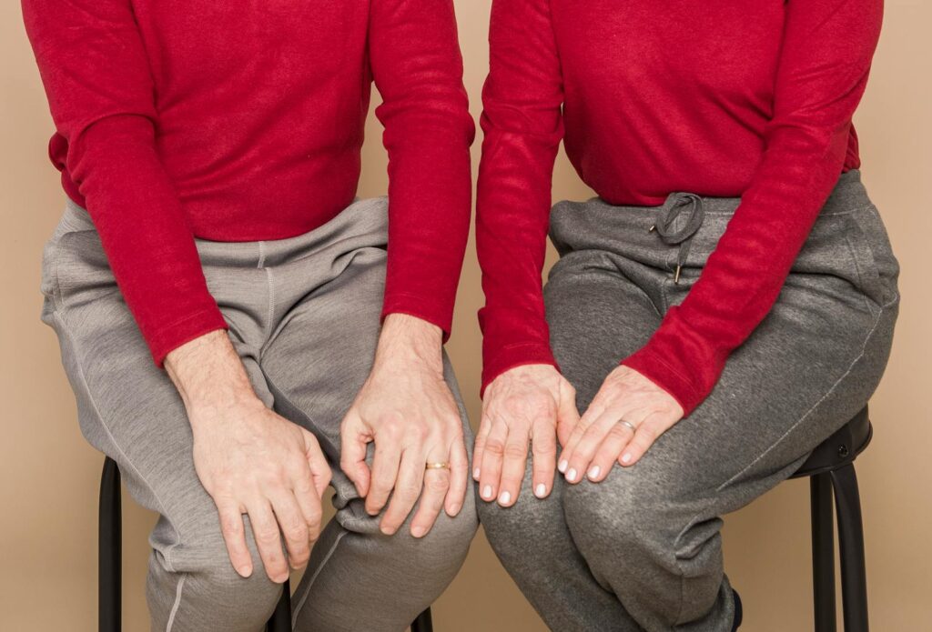 Crop unrecognizable mature man and woman in similar casual outfits sitting together on chairs with hands on knees against beige background