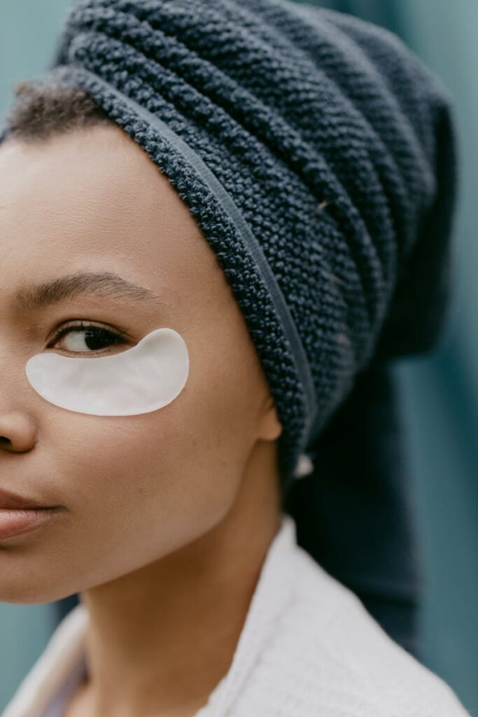 Close-up portrait of a woman with an eye patch and towel on head, showcasing skincare routine.