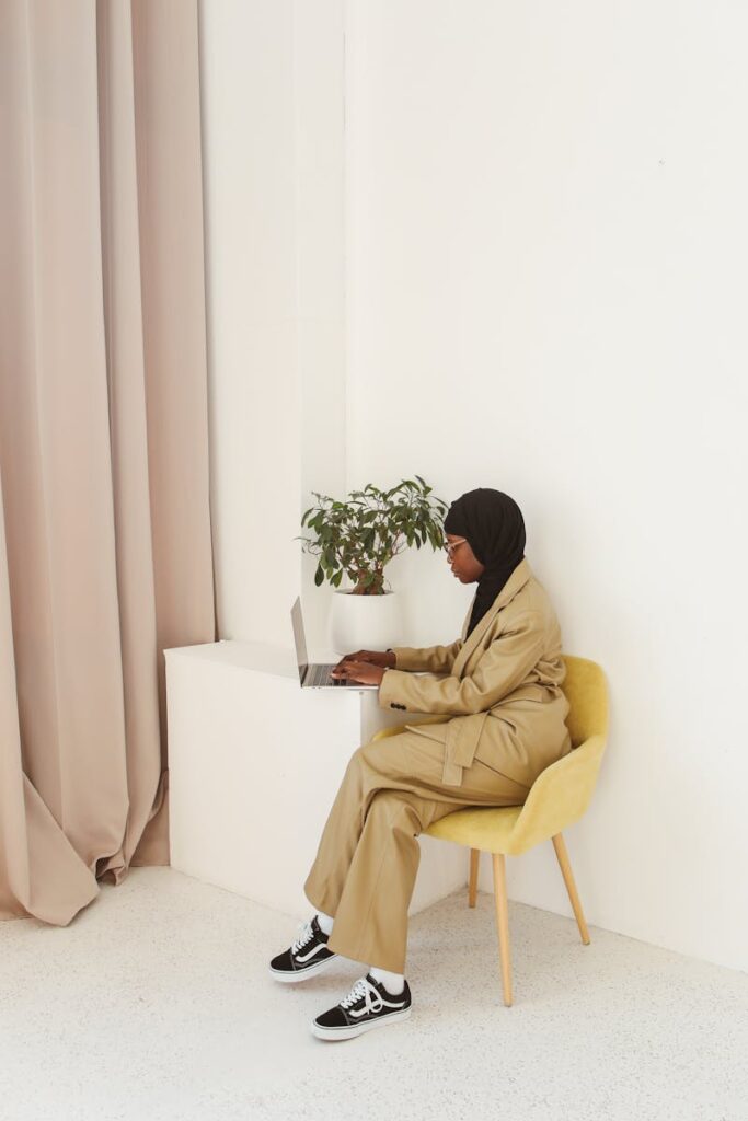 A Muslim woman in a hijab and stylish outfit works on a laptop indoors, exuding elegance and focus.