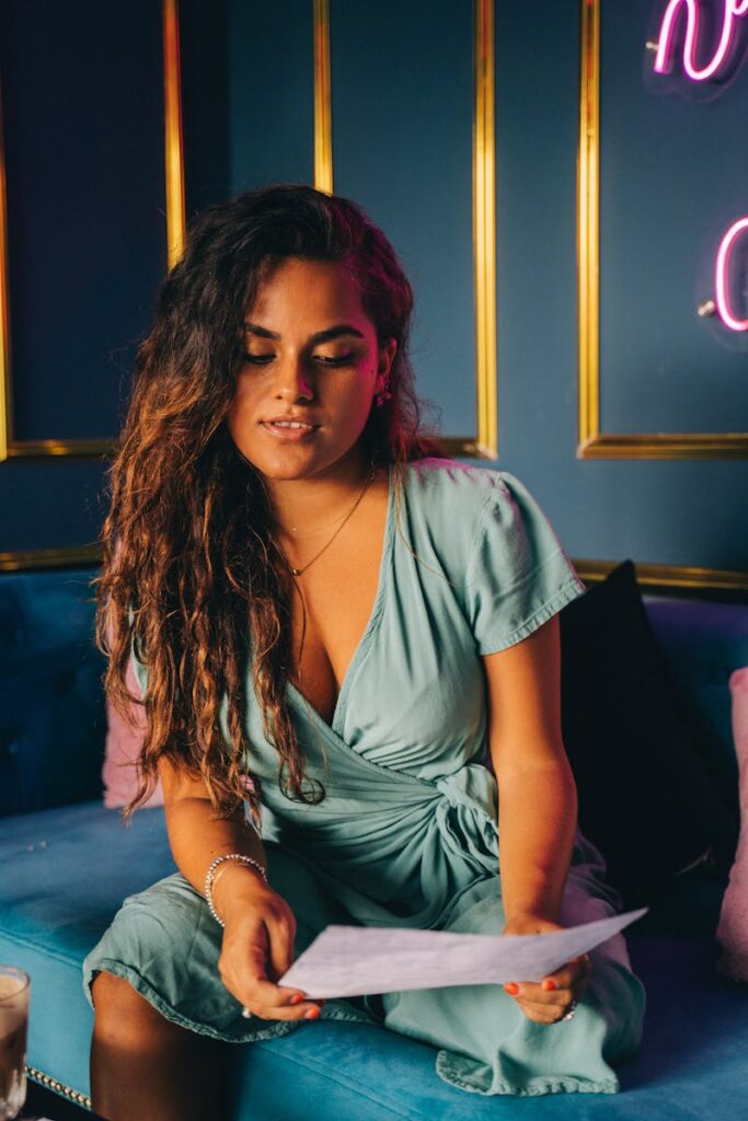 A woman in a teal dress sits on a couch reading a paper in a stylish room with neon lights.