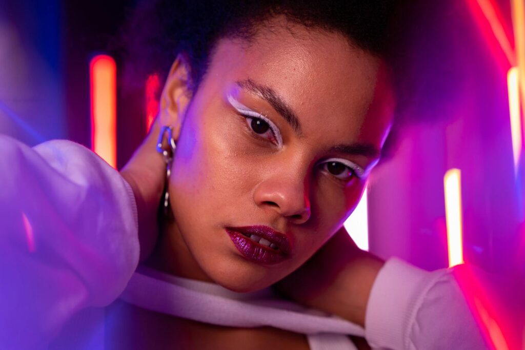 A striking portrait of a woman illuminated by neon lights, showcasing bold makeup and a serious expression.