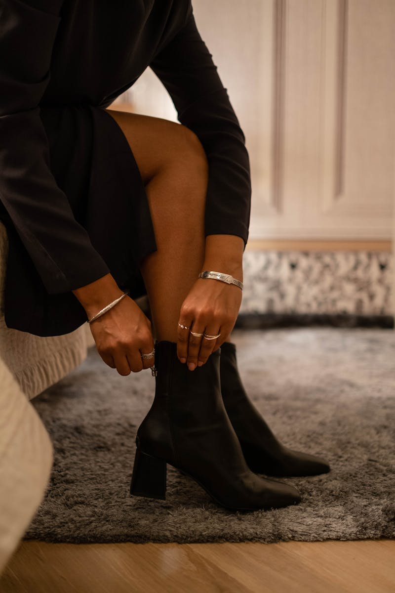 Fashionable woman putting on black ankle boots, showcasing accessories indoors.