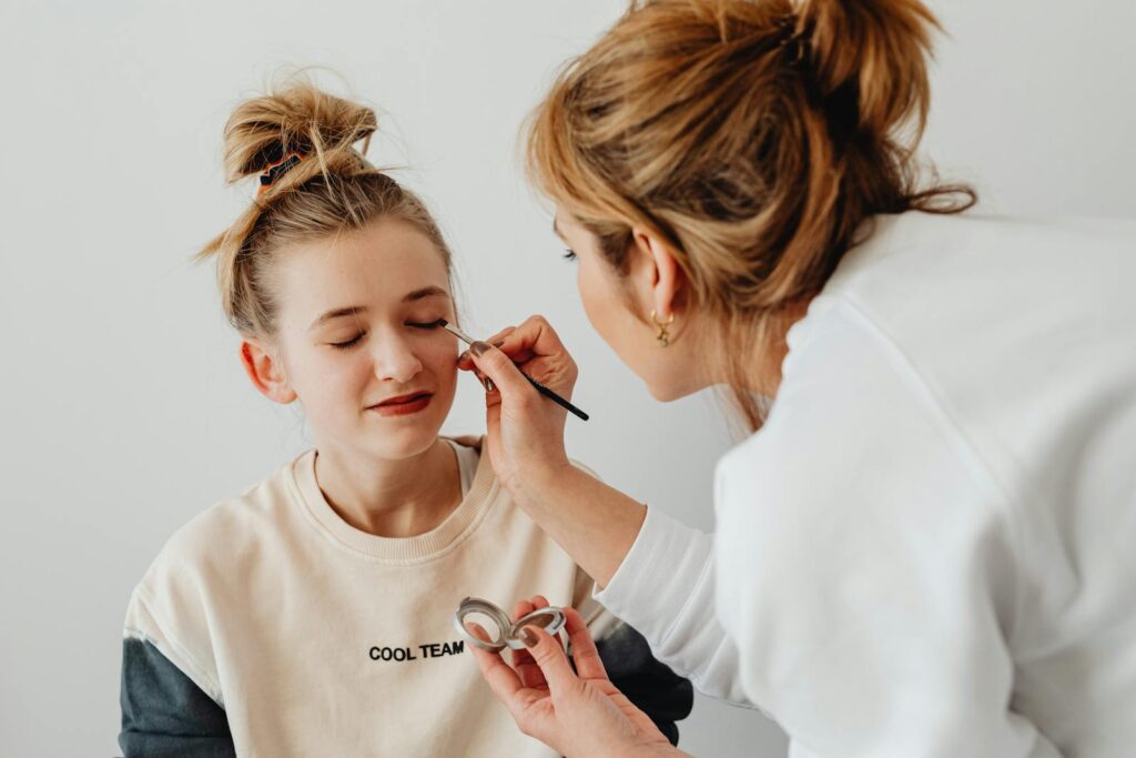 A mother is carefully applying eyeshadow to her daughter's closed eyes indoors.