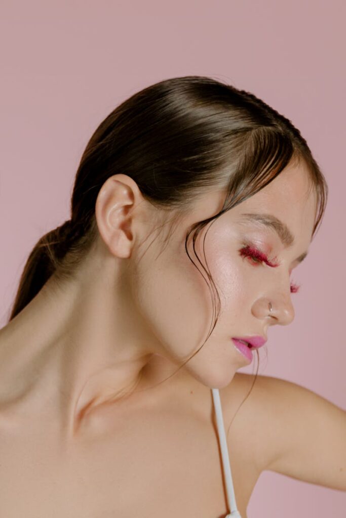 Close-up portrait of a woman with bold eye makeup and a nose piercing, set against a soft pink background.