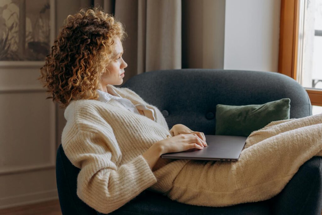 A woman with curly hair lounges on a sofa holding a laptop, enjoying a peaceful indoor moment.