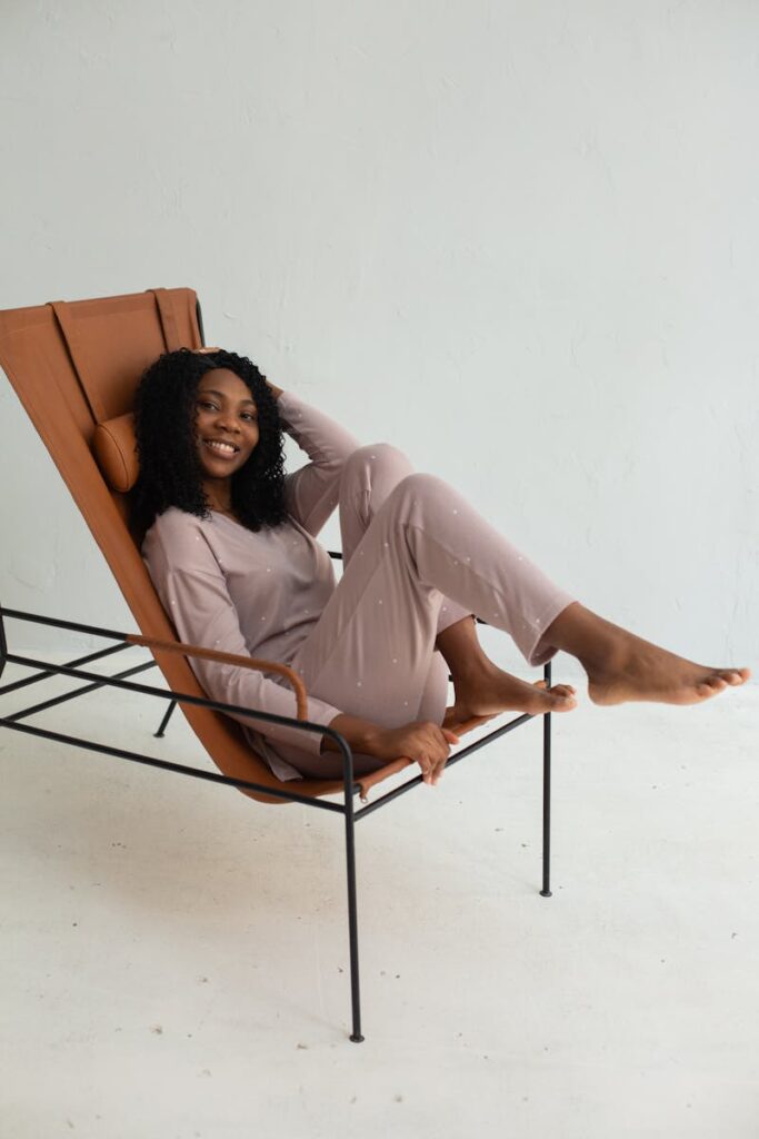 African American woman in loungewear smiling while sitting in a modern chair against a white background.