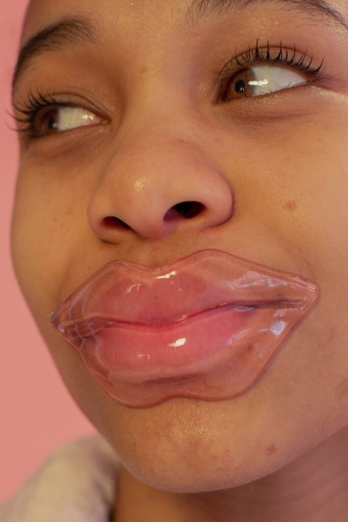 Close-up portrait of a young woman using a hydrating lip mask against a pink background.