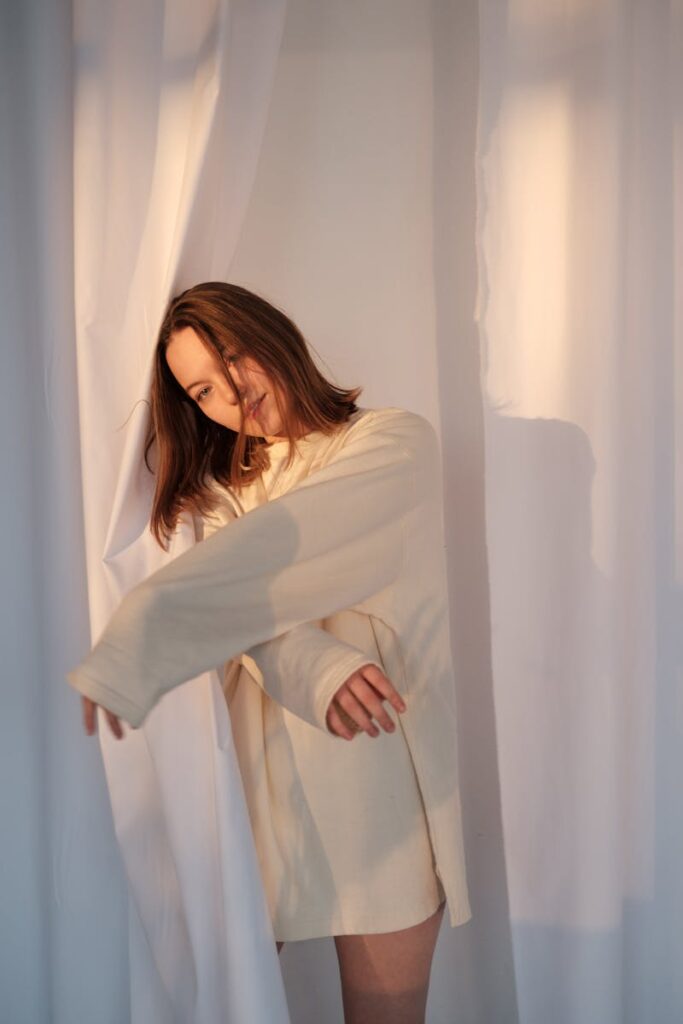 Young woman in sweatshirt leaning on white curtain and looking at camera in light room