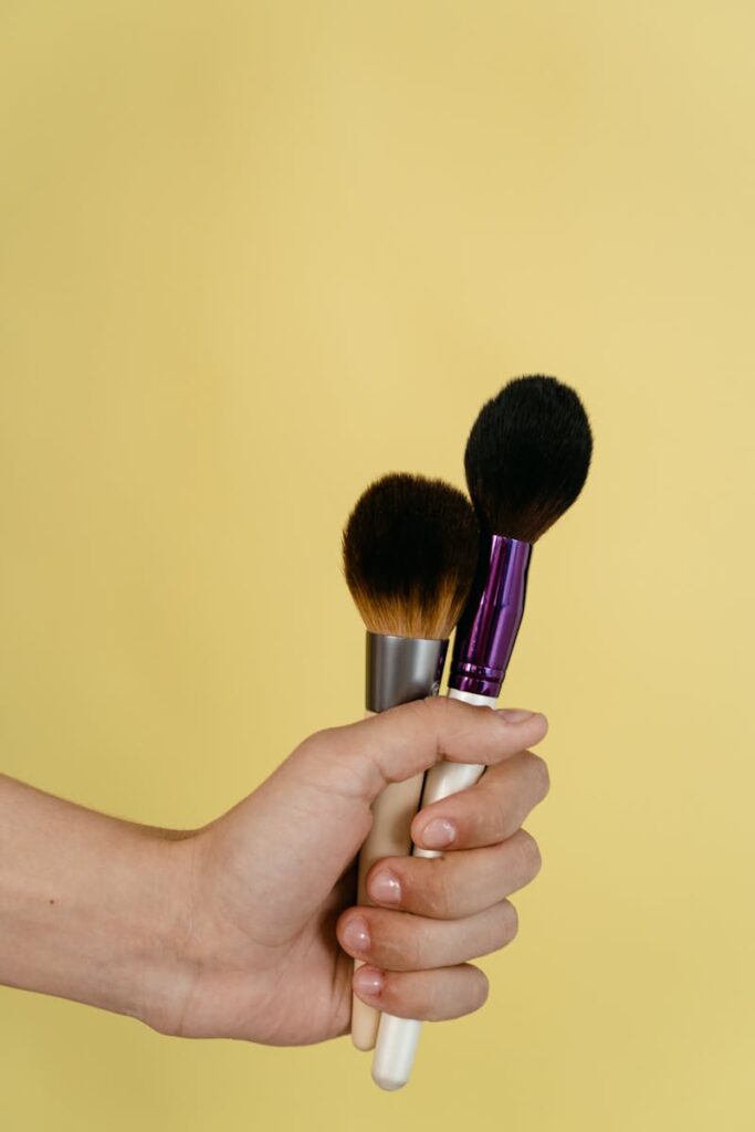 A hand holding two makeup brushes against a pale yellow background.