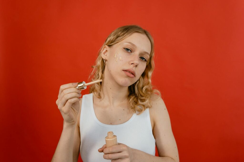 A young woman in a tank top applies concealer to her face with a dropper against a red background.