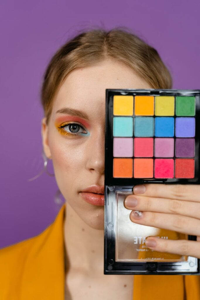 Close-up of a woman with colorful eye makeup holding a palette against a purple background.