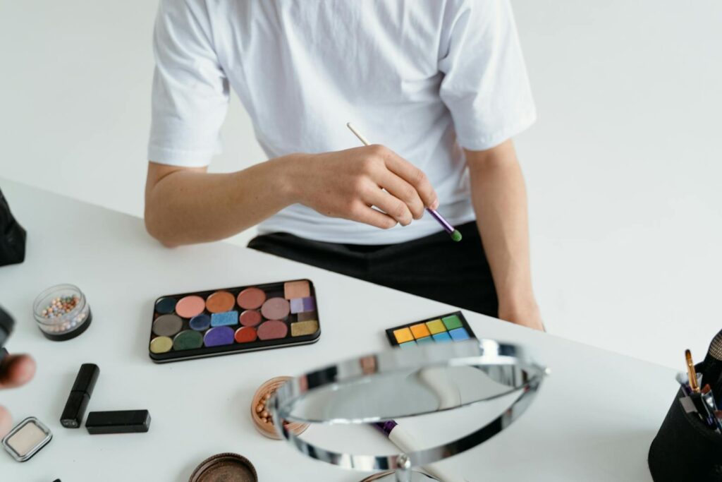 Close-up of a person applying makeup with a colorful eyeshadow palette and brush on a white table.