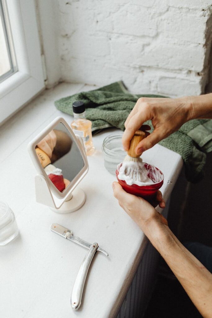 A vintage shaving setup with cream, a straight razor, and a mirror on a windowsill.