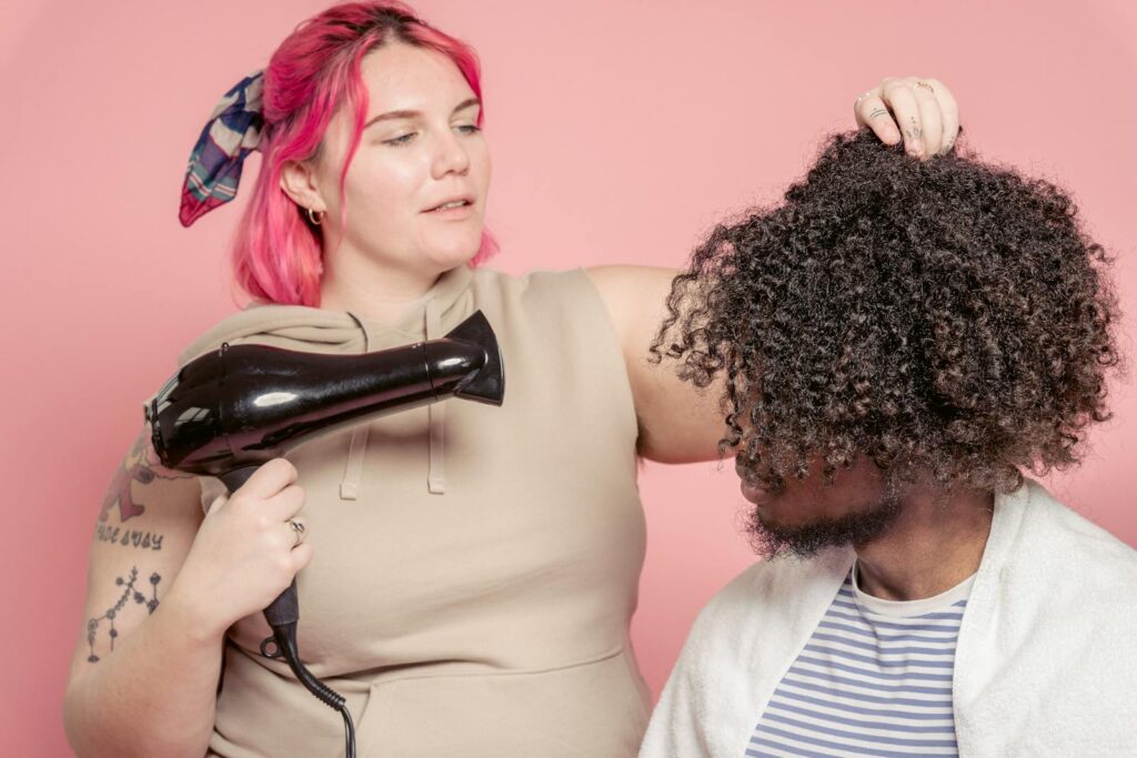 Female hairdresser with dyed hair drying hair of African American male model with Afro hairstyle against pink background