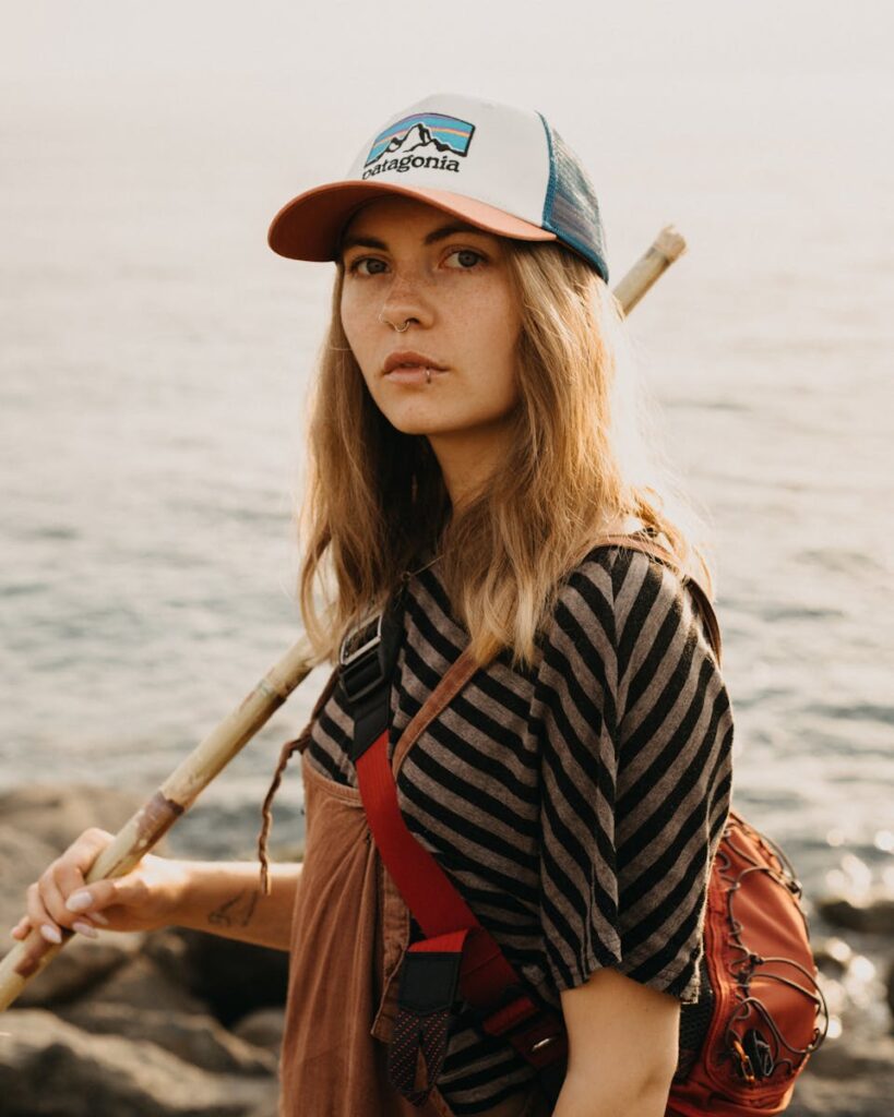 Serious female in cap with bag and bamboo stick looking at camera while standing on seashore