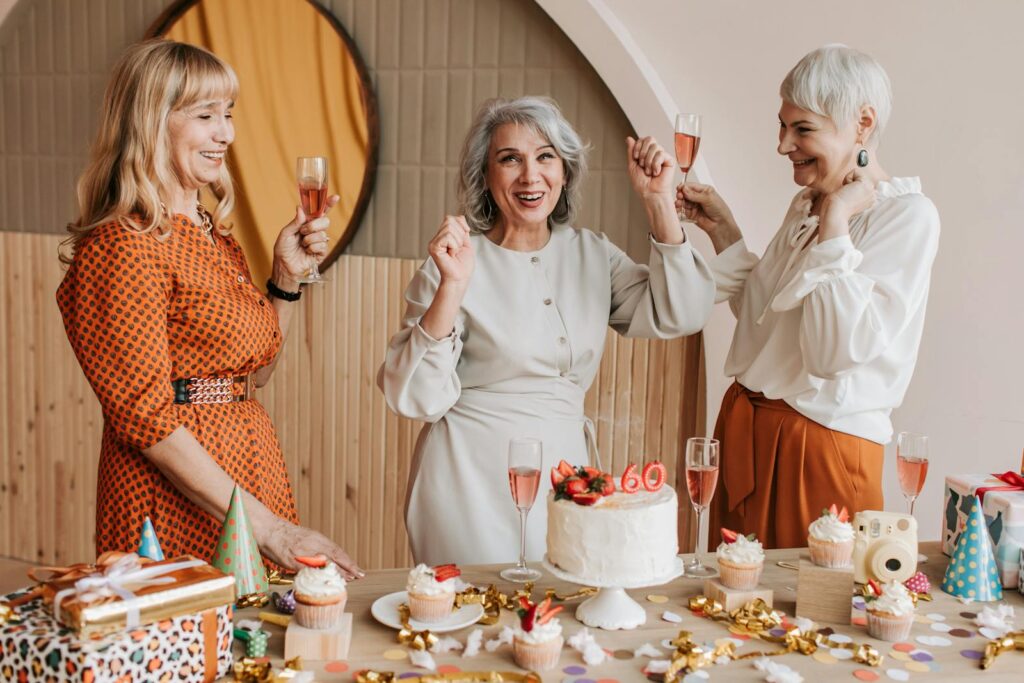 Three senior women celebrating a birthday with cake and champagne indoors.