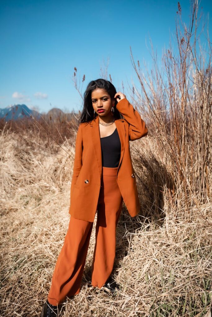Woman in brown blazer and trousers poses confidently in a sunny field.