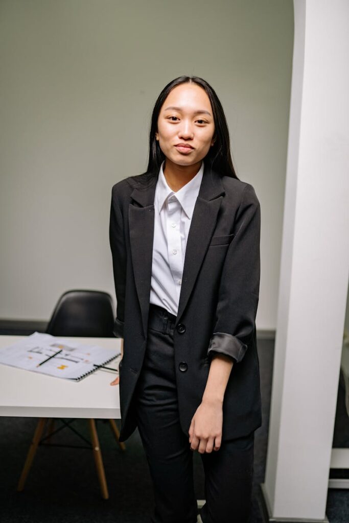 Asian woman in a formal black suit with white shirt standing confidently in an office setting.