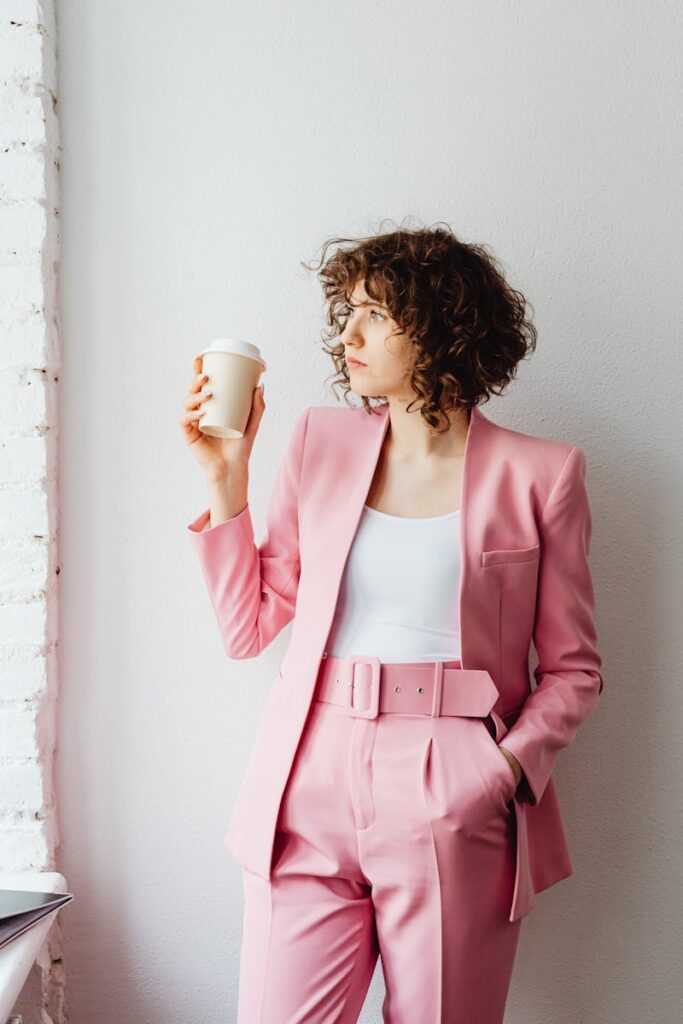 Businesswoman in a stylish pink suit holding a coffee cup, standing indoors with a thoughtful expression.