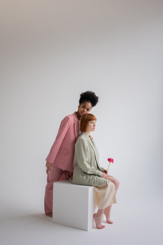 Full body of African American female in trendy suit standing behind ginger haired woman with flower sitting on cube against gray background