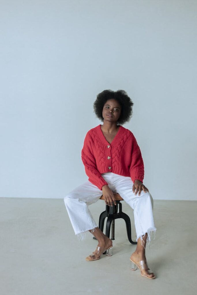 Fashionable woman in red sweater and white pants sitting on stool in a studio.