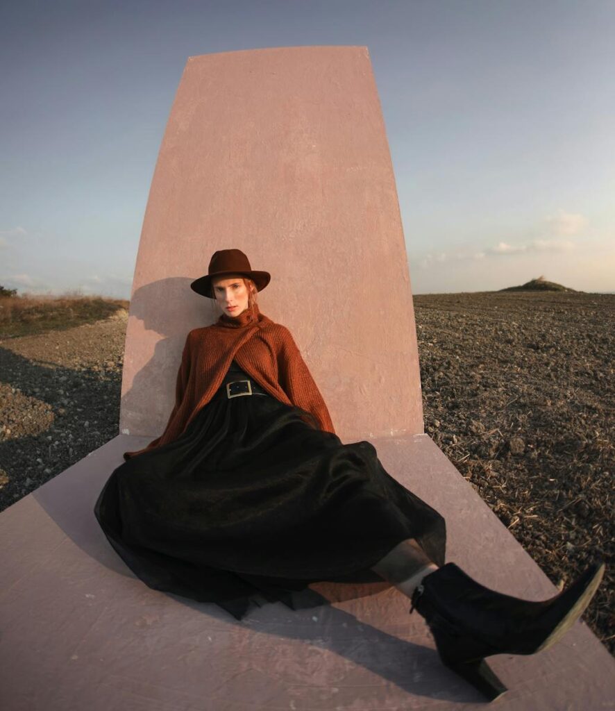 Full body of fashionable female in trendy outfit with hat looking at camera while sitting on decoration in woods with grassy field