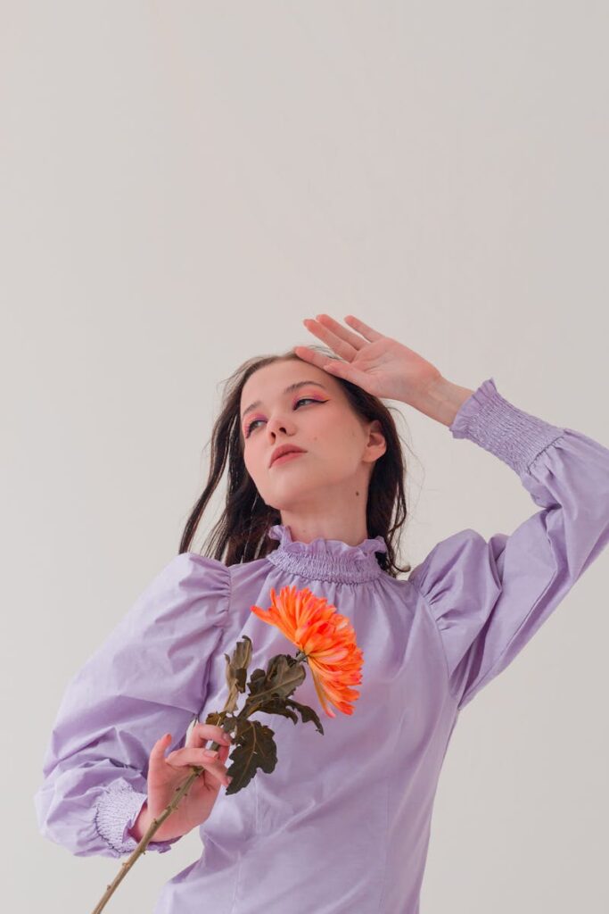 A fashionable woman in a lavender dress poses elegantly indoors holding a vibrant orange flower.