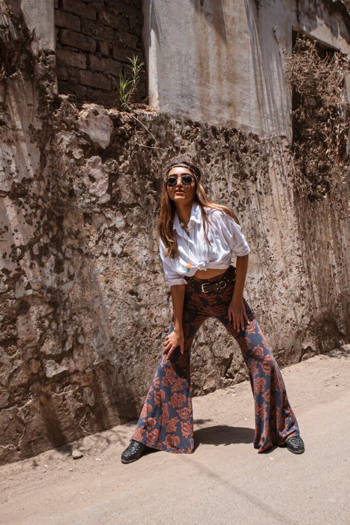 Fashion-forward woman poses in bell bottom pants against a gritty urban wall outdoors.