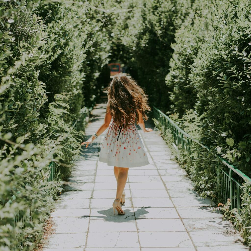 Back view of a woman in a sunlit park walking amidst lush green trees on a paved pathway.