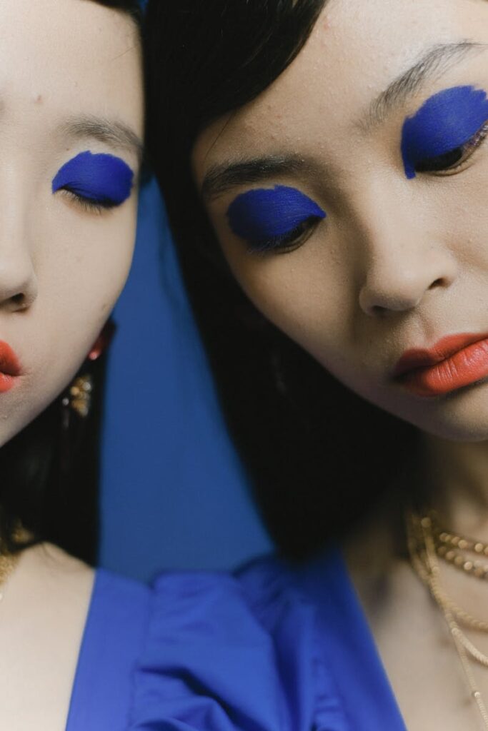 Artistic close-up of two women with striking blue eye shadow against a blue background.