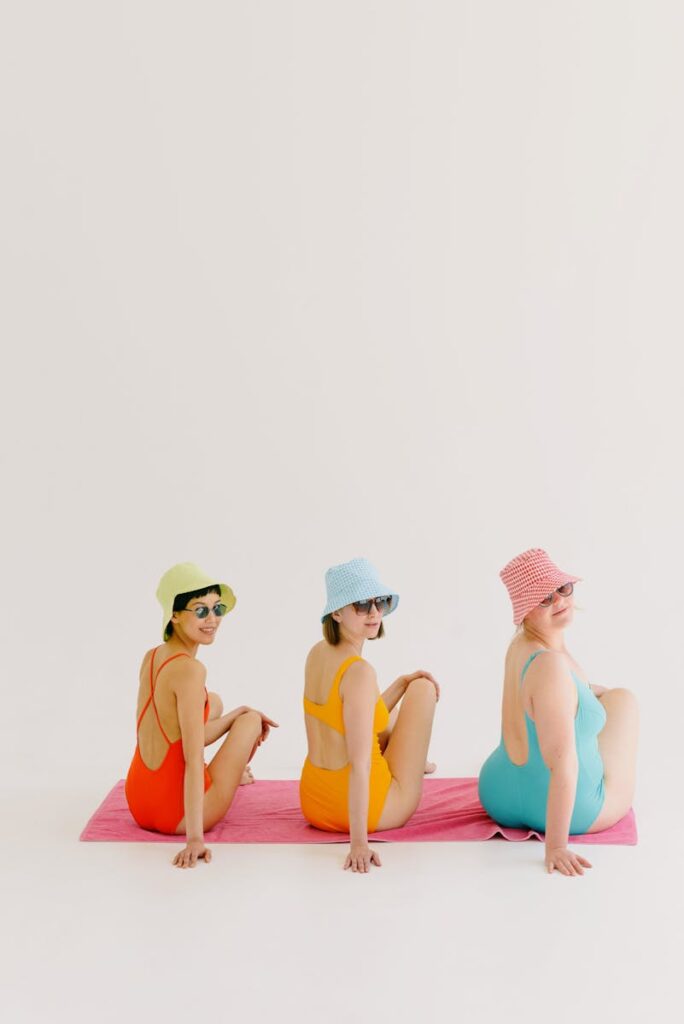 Three young women in colorful swimsuits posing sitting on a towel with bucket hats and sunglasses.