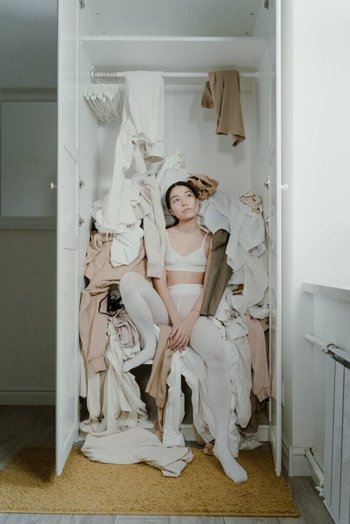 A contemplative woman sits surrounded by clothes in a messy wardrobe, evoking themes of consumerism and clutter.