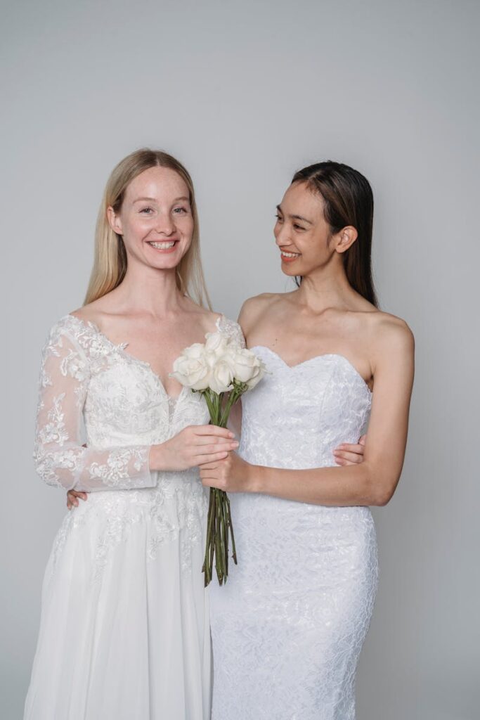 Two brides wearing elegant dresses with white bouquets, smiling in a studio setting.