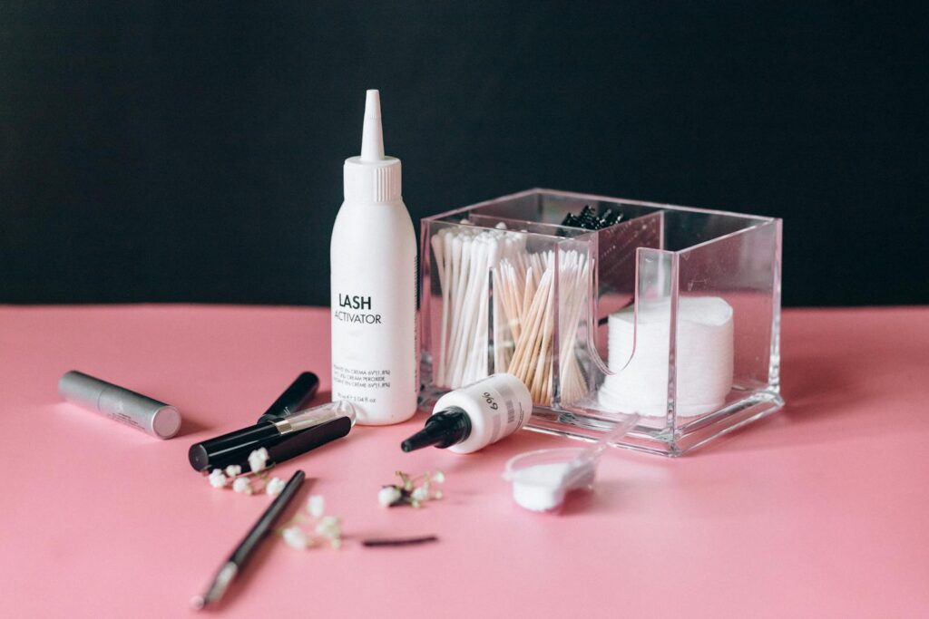 A selection of makeup tools and skincare products arranged on a pink surface, including lash activator and cotton pads.