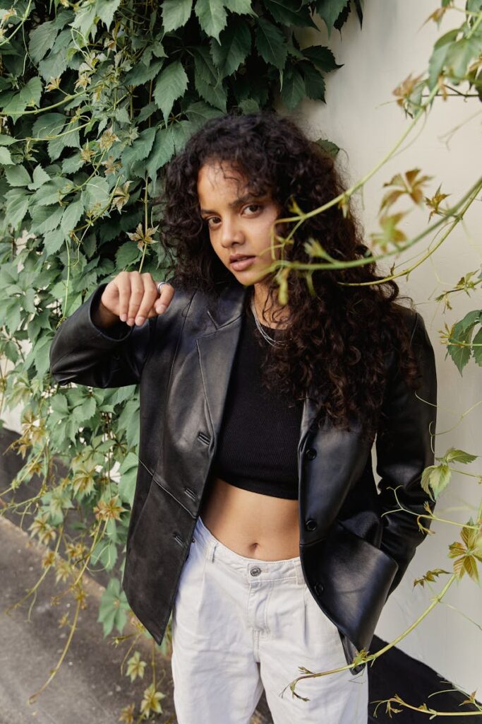 A stylish woman in a leather blazer poses in an urban setting with green vines.