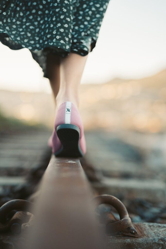 A woman in pink shoes and a floral dress balances on a train track as the sun sets.
