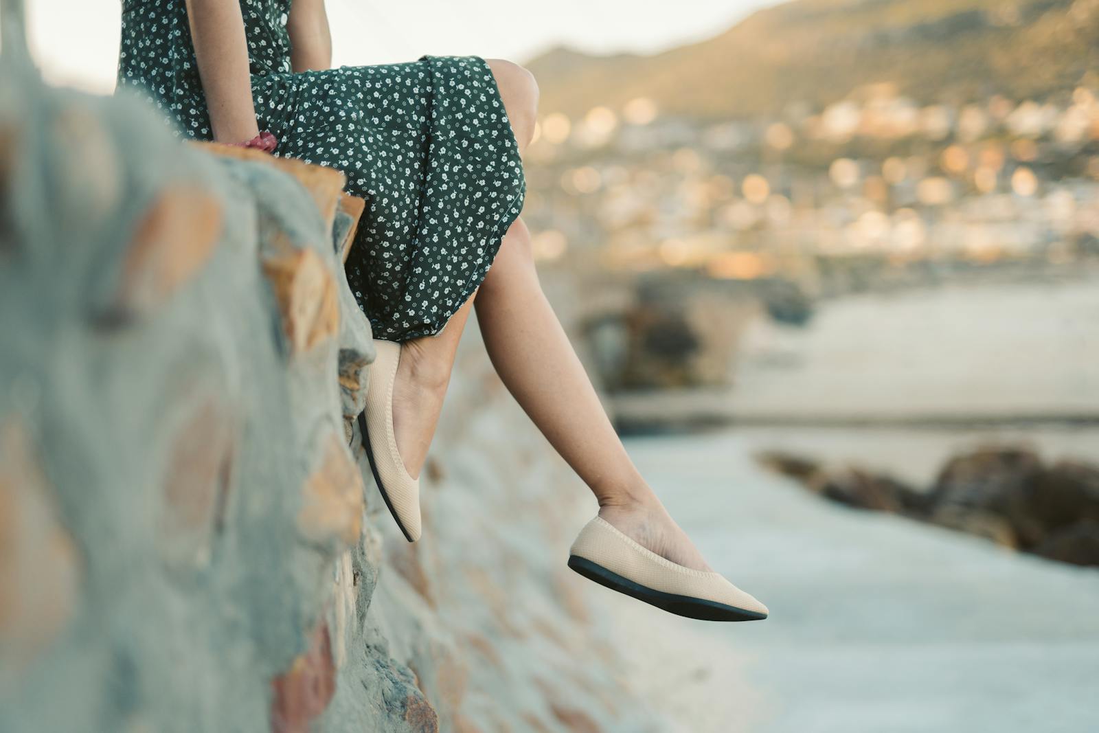 A woman sitting by the beach wearing a floral dress and flats, enjoying a summer sunset.