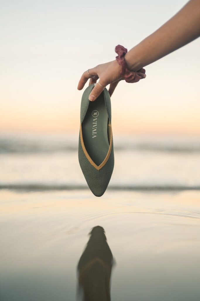 A woman's hand holding a fashionable shoe over water during sunset.