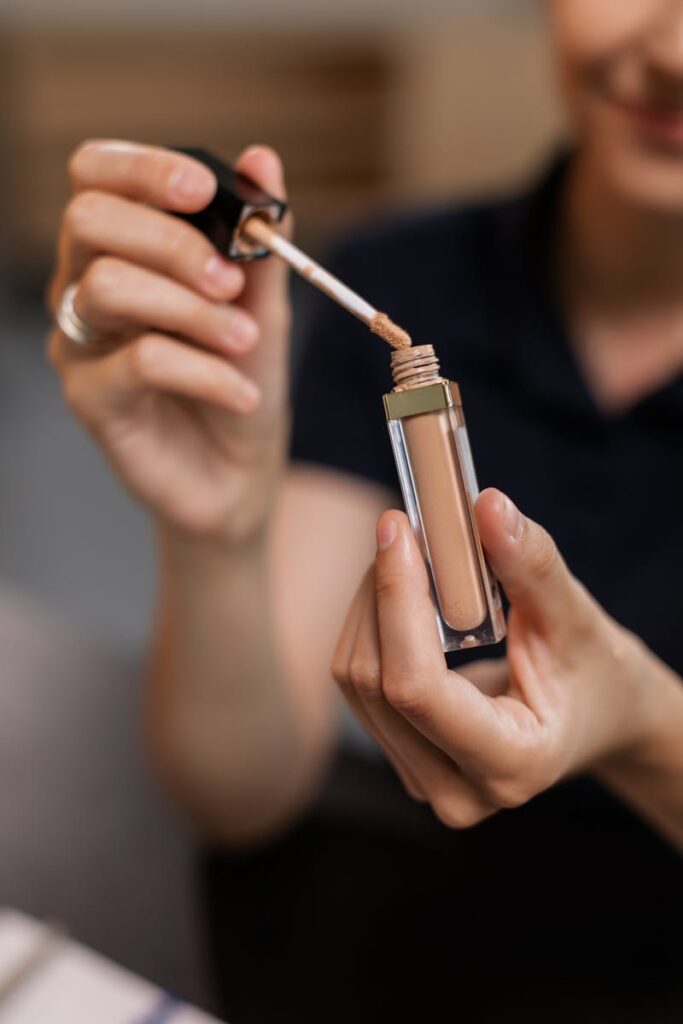 Close-up of a woman's hands holding and applying liquid concealer indoors.