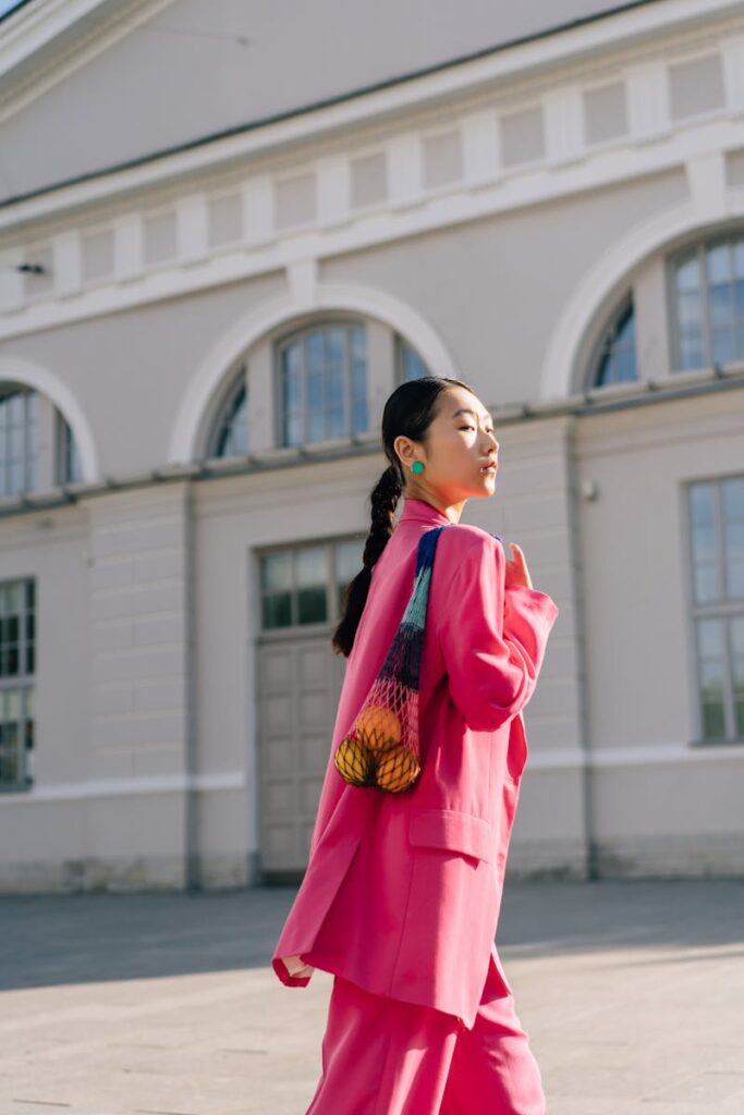 Stylish woman in vibrant pink suit walking outdoors, eco-friendly net bag with oranges.