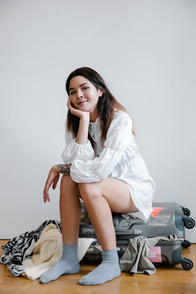 Smiling woman in white dress sitting on luggage amidst casual clothes, indoors.