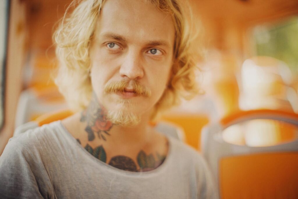 Close-up portrait of a blonde man with tattoos sitting in a brightly lit environment.